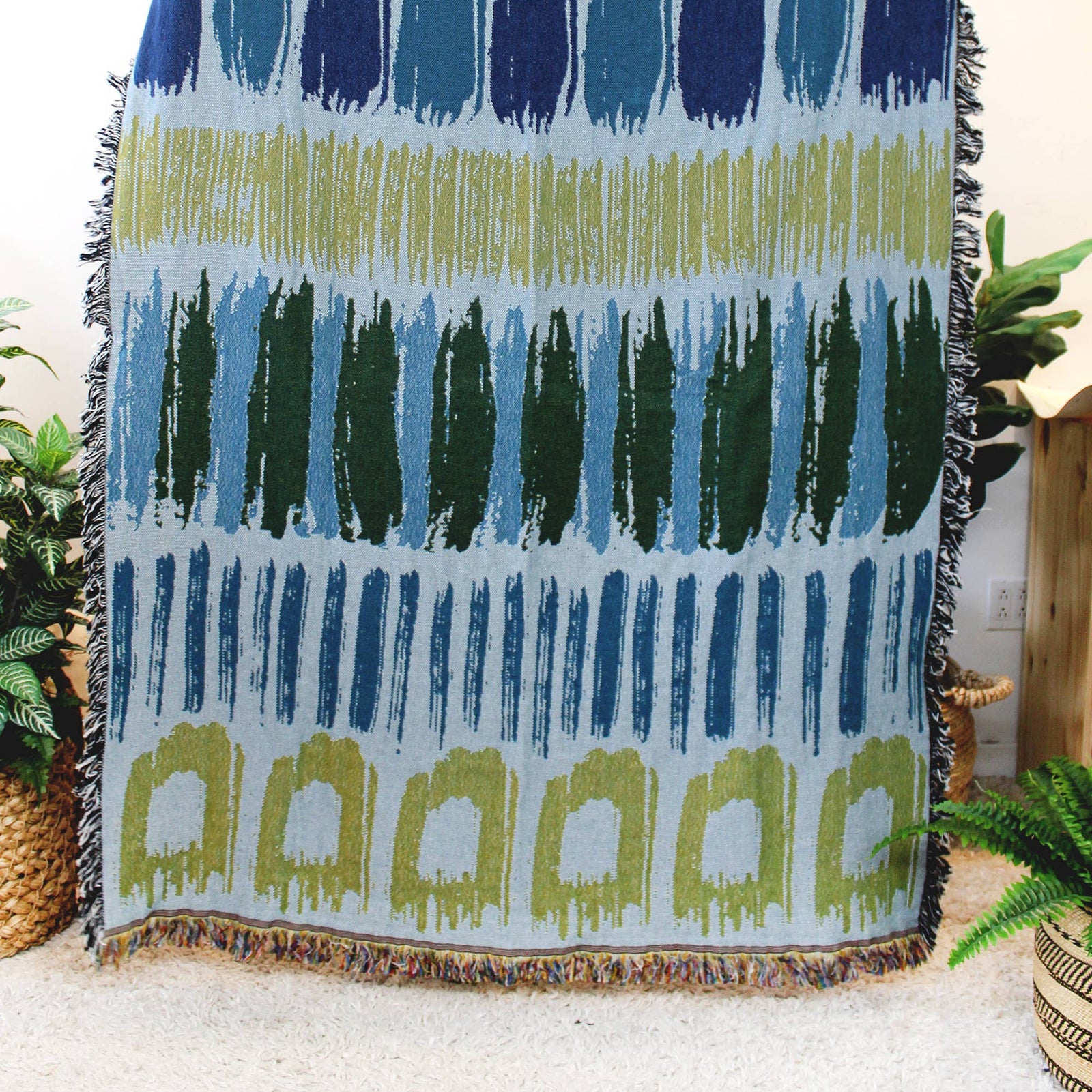 A woven throw blanket with a geometric pattern in shades of blue, green, and olive on a light background, featuring black fringe on all sides, displayed in a home setting.