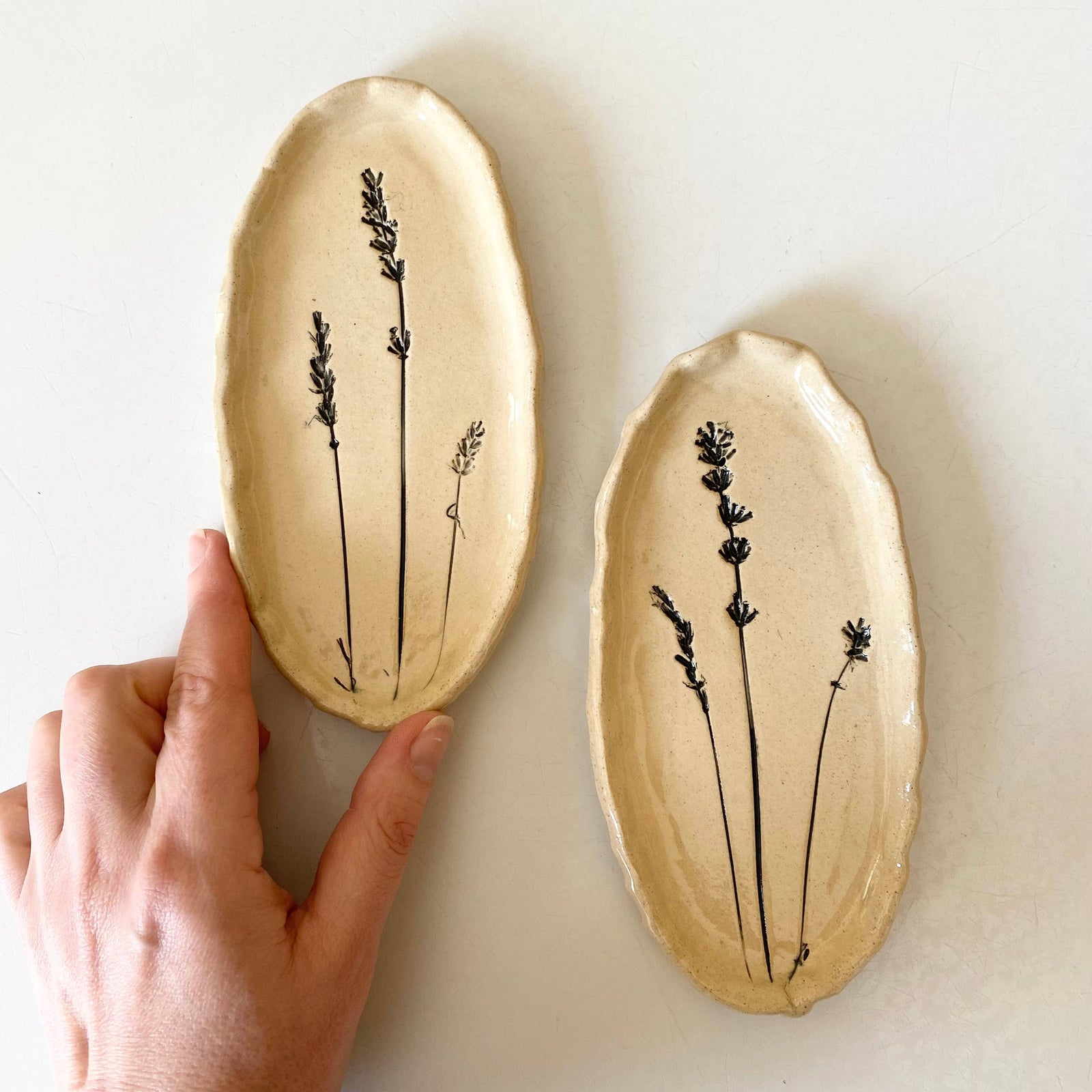 Two oval ceramic plates with black plant designs on a white background, one held by a hand.