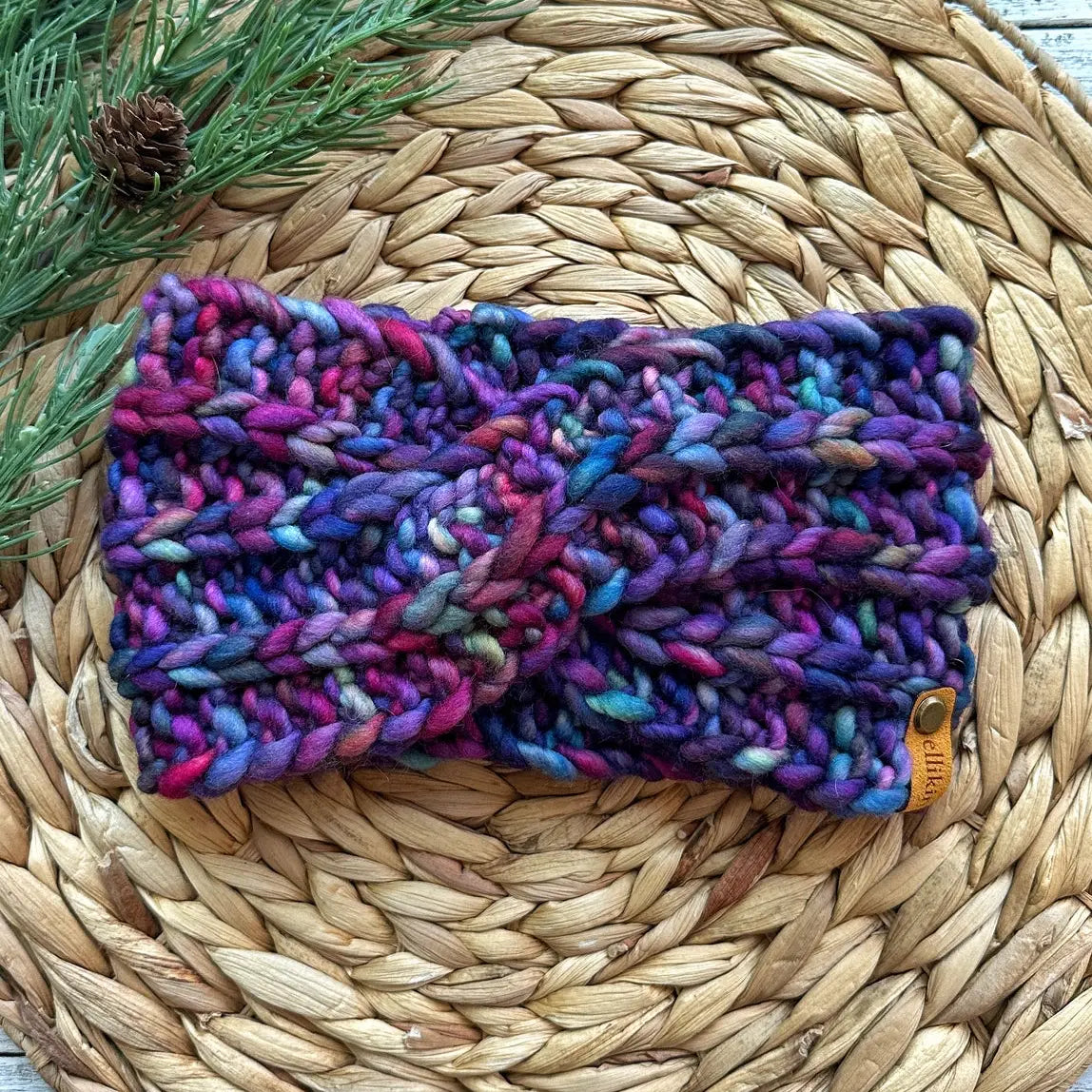Multicolored knitted headband on a woven mat with a pine cone and branch.