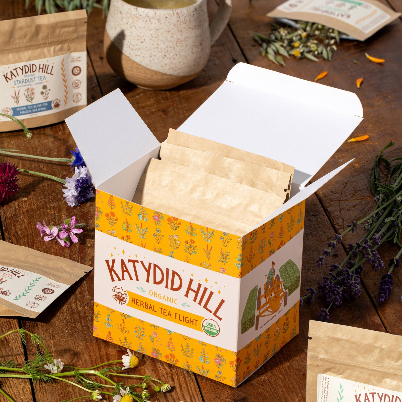 A box of Katydid Hill organic herbal tea  flight samples, with individual tea bags displayed in front and a mug in the background, surrounded by decorative flowers.