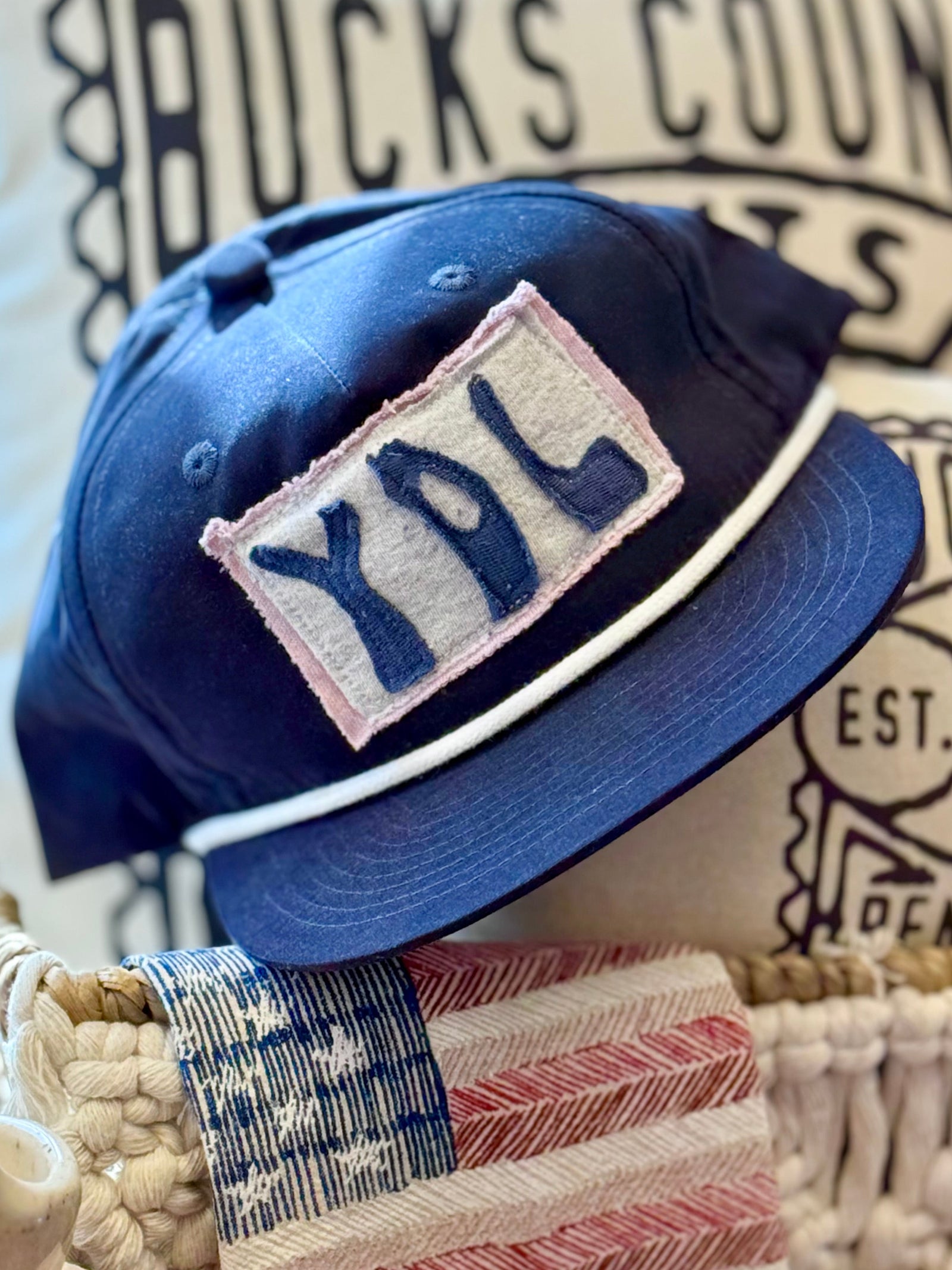 A navy blue baseball cap with a white embroidered patch reading 'YDL' (three letter borough code for Yardley PA 19067) in navy on a light pinkish-beige background, styled on a woven basket with a rustic decorative pillow and flag towel 