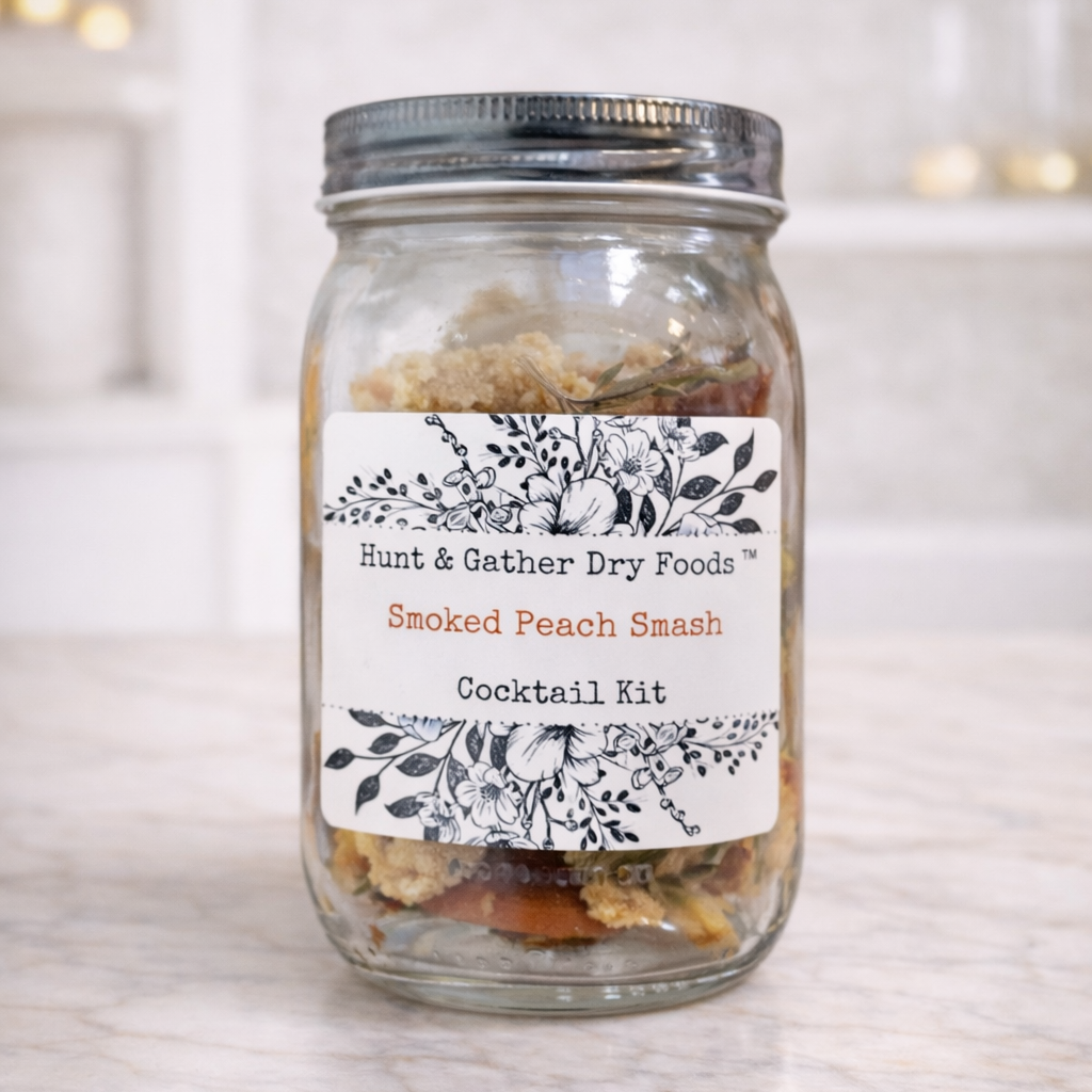 A clear glass mason jar with a metal lid and a white decorative label reading 'Hunt & Gather Dry Foods Smoked Peach Smash Cocktail Kit,' set on a light marble surface with a softly blurred kitchen background.