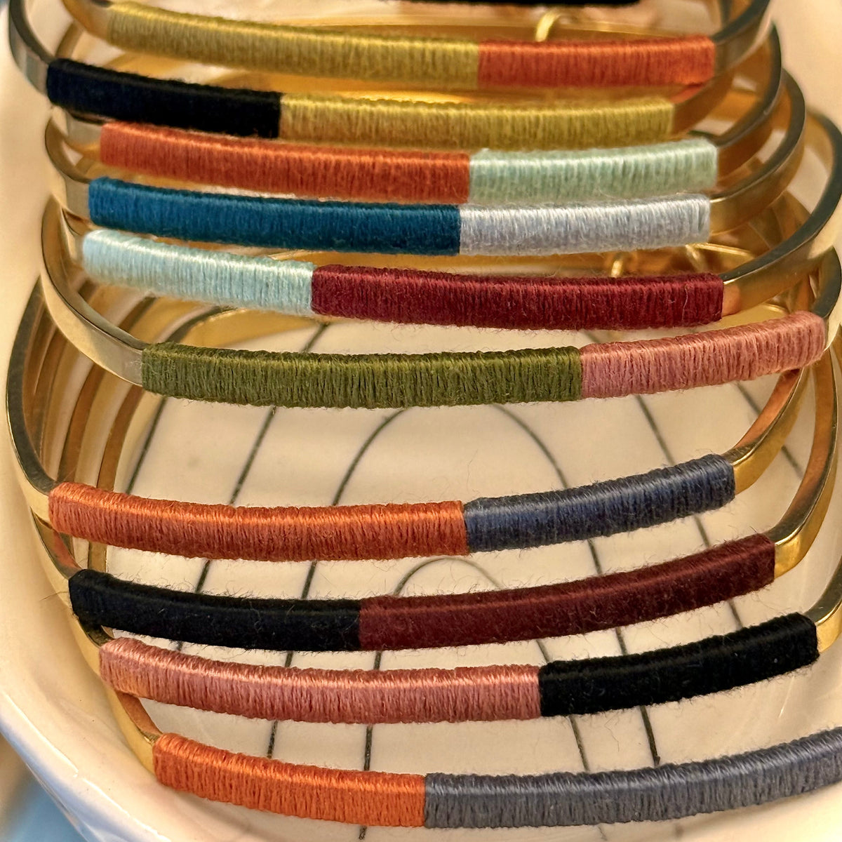 A stack of gold-plated bangle bracelets with colorful striped woven bands, displayed on a white plate.
