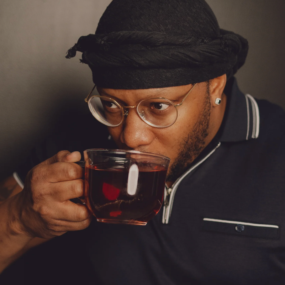 Person wearing a black cap and glasses, holding a glass mug of herbal tea.