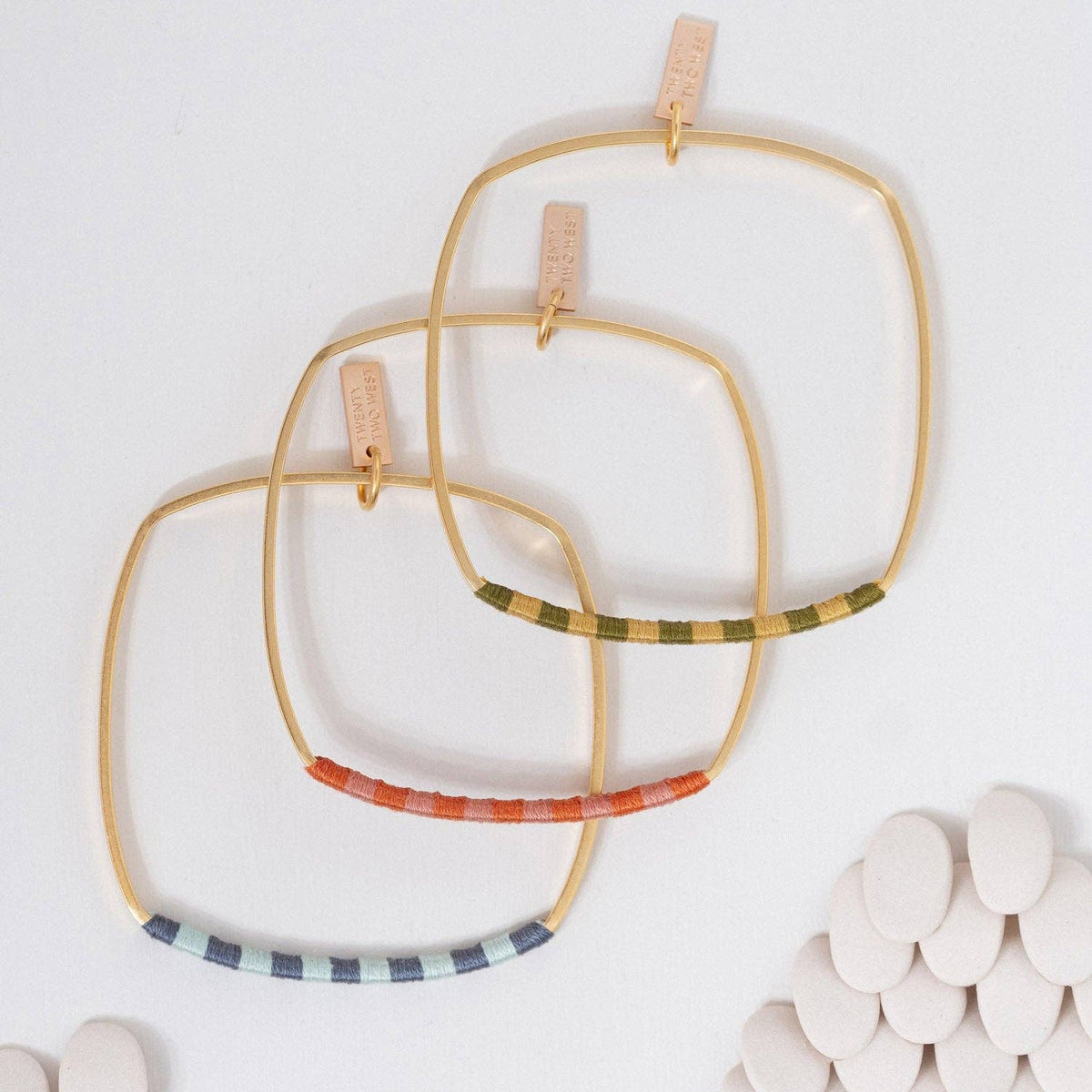 Three large square gold hoop bangles with colorful striped accents, displayed on a white background with decorative white stones.